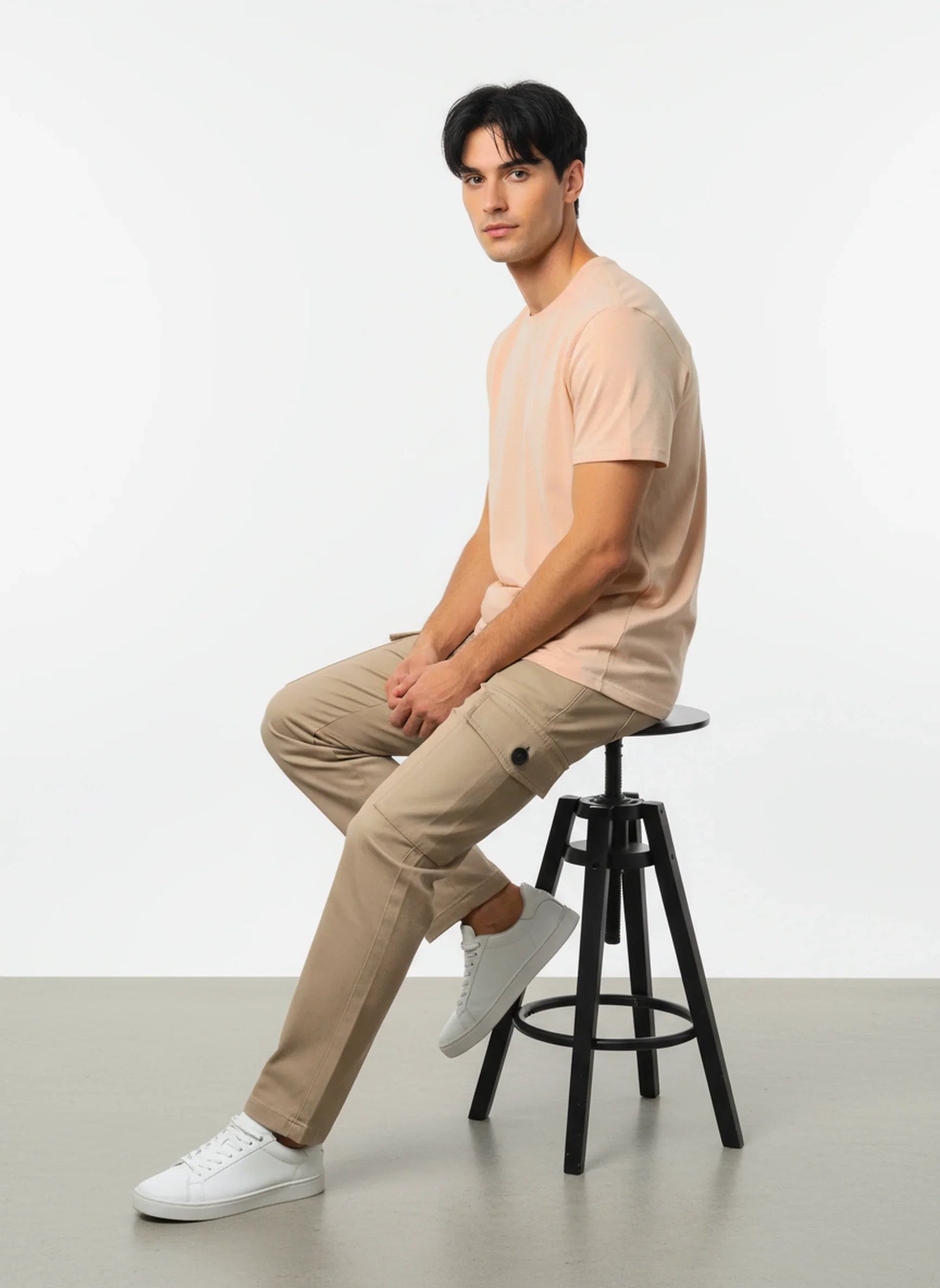Man sitting on black stool wearing peach t-shirt, beige cargo pants, and white sneakers on gray floor