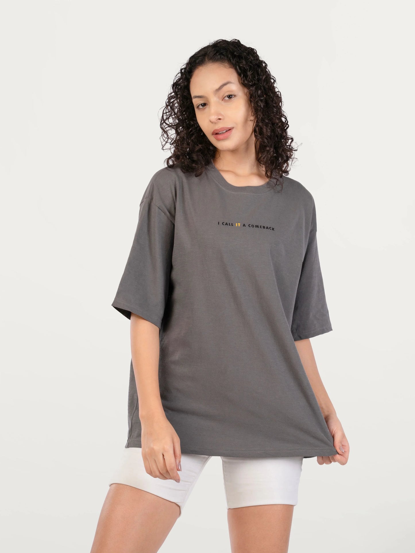 Young woman with curly hair wearing oversized gray T-shirt with text I CALL IT A COMEBACK and white shorts