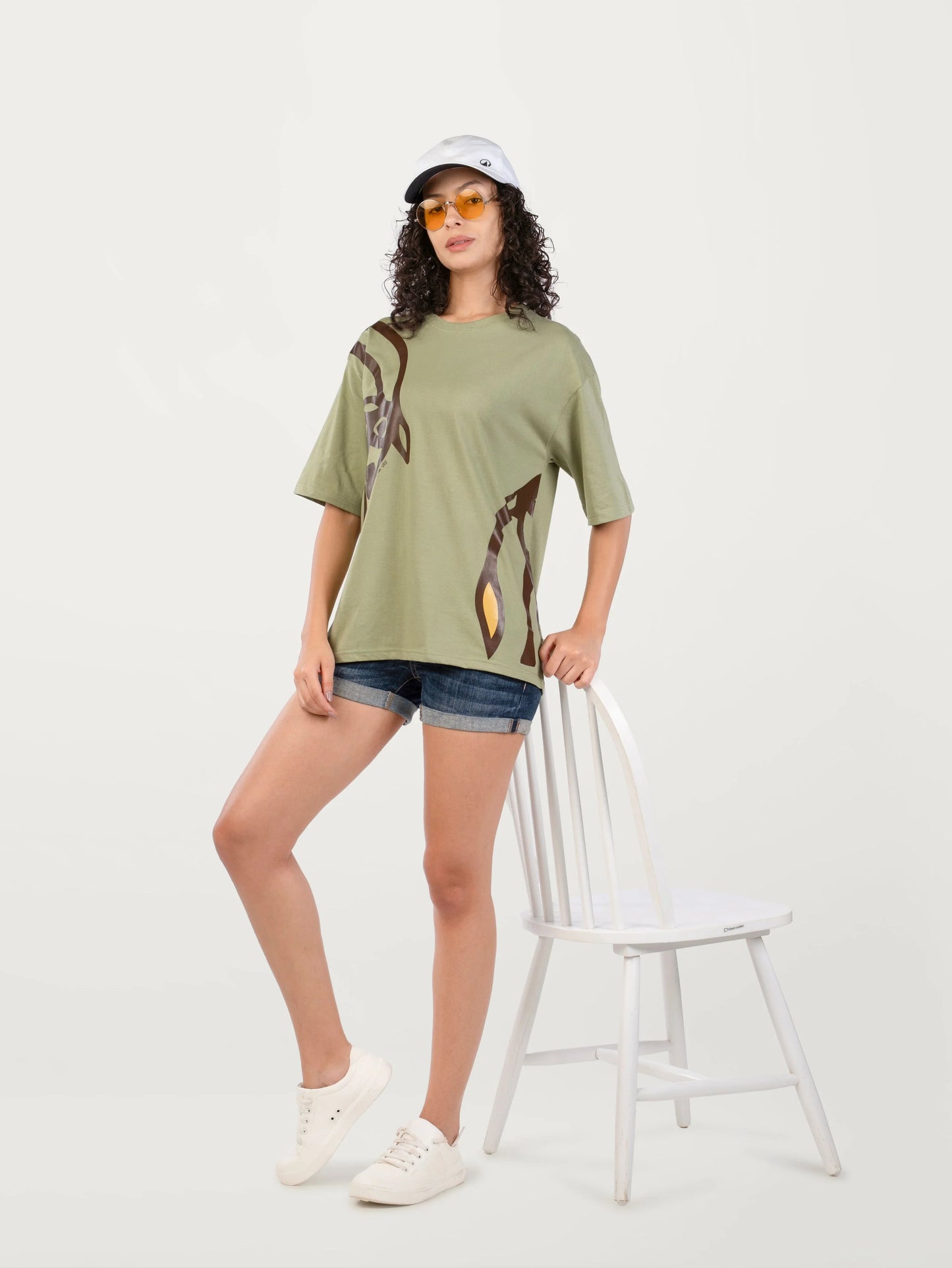 Woman wearing olive green graphic t-shirt, denim shorts, white sneakers, white cap, and yellow sunglasses leaning on white chair