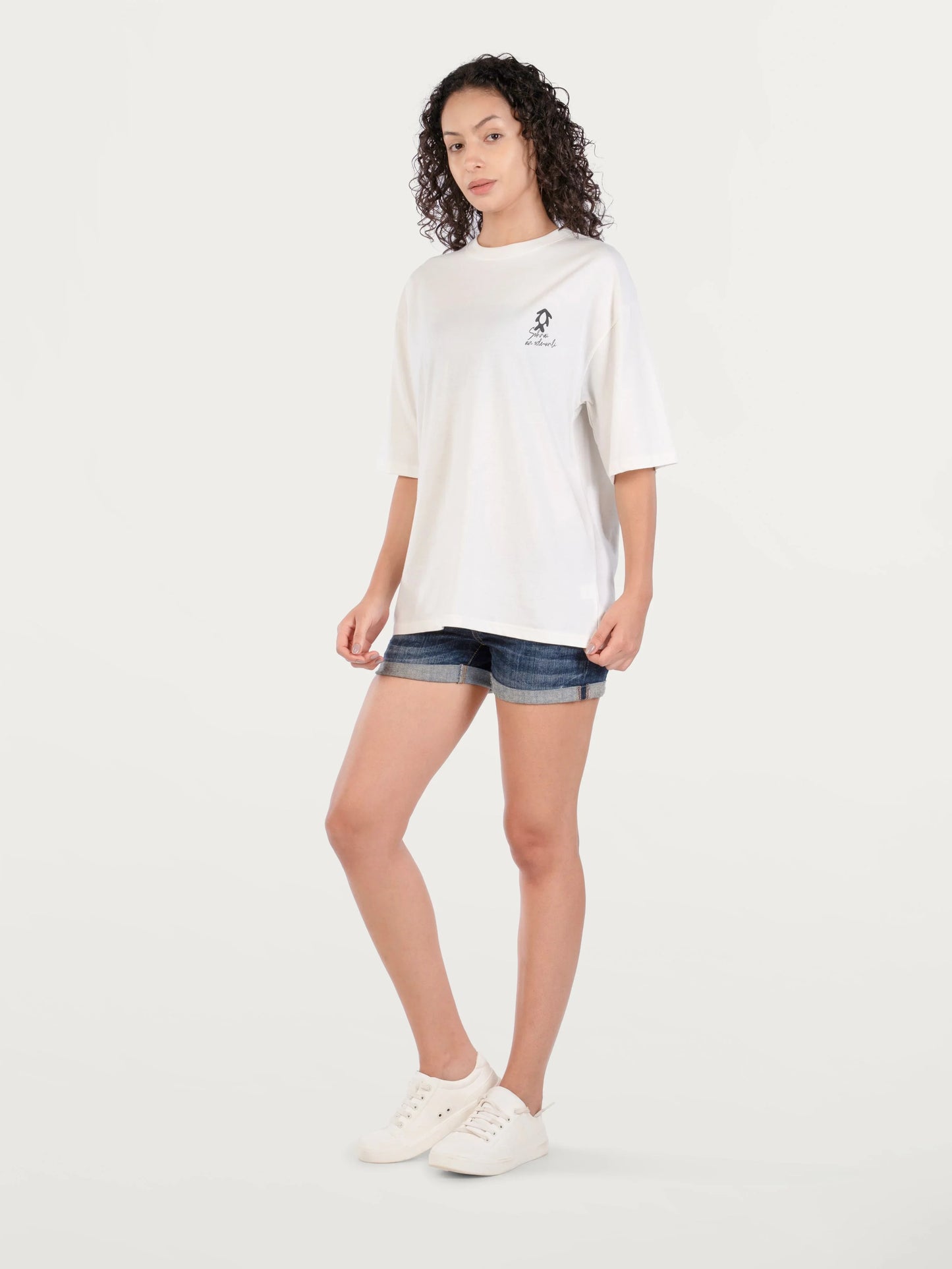 Woman in white graphic T-shirt, denim shorts, and white sneakers on plain background