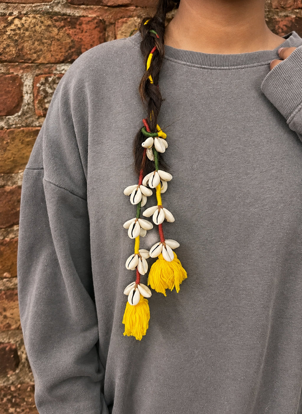 A close-up of a person wearing a gray sweatshirt, showing a long braided braid adorned with white cowrie shells and bright yellow tassels.