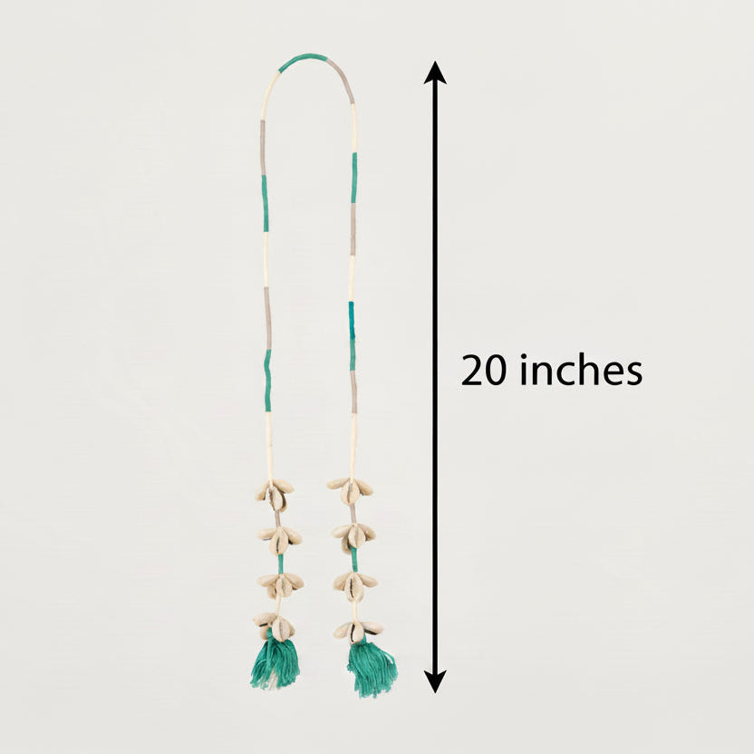 A long bohemian-style necklace featuring a cream and green beaded cord, shell-like beads, and two green tassels at the bottom, displayed against a solid off-white background.