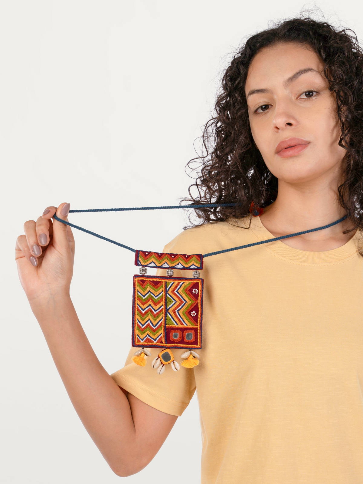 A woman with curly dark hair wearing a yellow t-shirt holds up a small rectangular woven pouch with a dark blue rope strap. The pouch features a vibrant geometric pattern in red, yellow, green, and white, with small yellow tassels hanging from the bottom.
