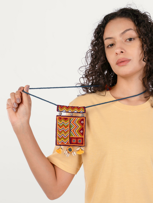 A woman with curly dark hair wearing a yellow t-shirt holds up a small rectangular woven pouch with a dark blue rope strap. The pouch features a vibrant geometric pattern in red, yellow, green, and white, with small yellow tassels hanging from the bottom.